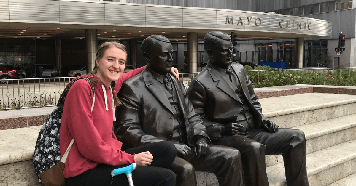 Kaley, with braids and a pink hoodie, posing with the statues of the Mayo brothers in front of the Mayo Clinic building.