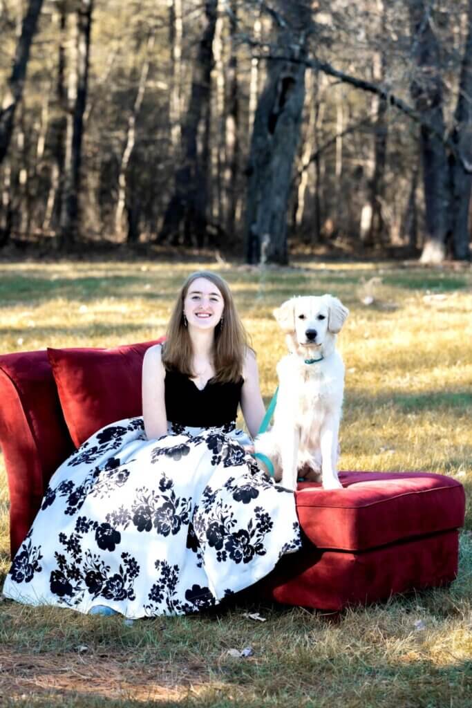 Kaley, wearing a black and white floral dress, sitting with Phyllo the golden retriever on a red couch in the middle of a grassy and wooded area.